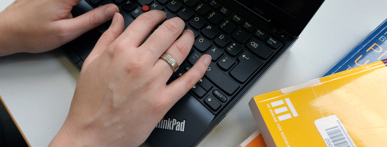 Eine Studentin tippt an einem Laptop und gelbe Bücher legen daneben.